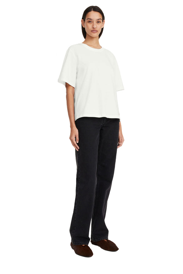 Front view of a woman wearing By Malene Birger t-shirt in white, paired with black trousers and a pair of brown flats, shown against a plain white background.