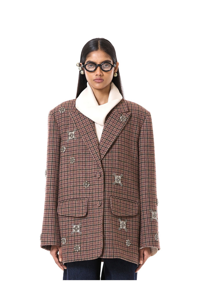 A model wearing a brown and beige plaid double-breasted blazer with embroidered details, a white turtleneck underneath, and dark jeans, photographed against a solid white background.